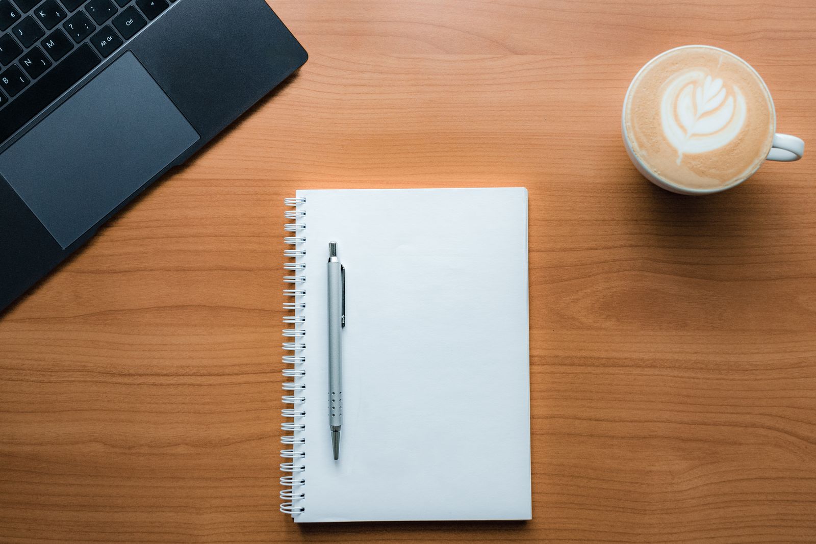Aufgeschlagenes Notizbuch, Laptop und eine Tasse Kaffee auf einem Holztisch, von oben fotografiert